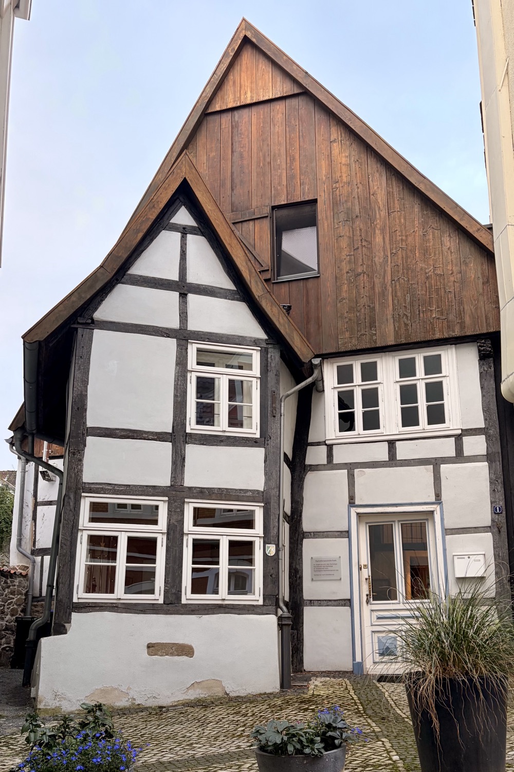 Außenfassade des denkmalgeschützten Fachwerkhauses Schießhofstraße 1 mit spitzem Giebel, Fachwerk, blau gerahmter Eingangstür und Pflanzkübeln auf dem Kopfsteinpflaster davor.