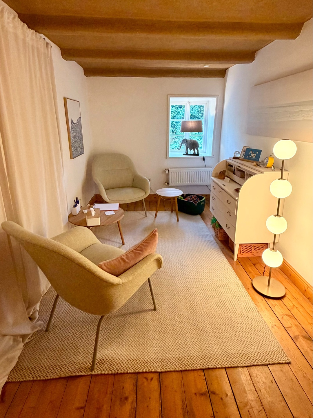 Warm beleuchteter Behandlungsraum unter freiliegenden Holzbalken mit zwei Sesseln in Salbeigrün, zartrosa Kissen, hellem Teppich, einem Sekretär und einer Kugelstehlampe.